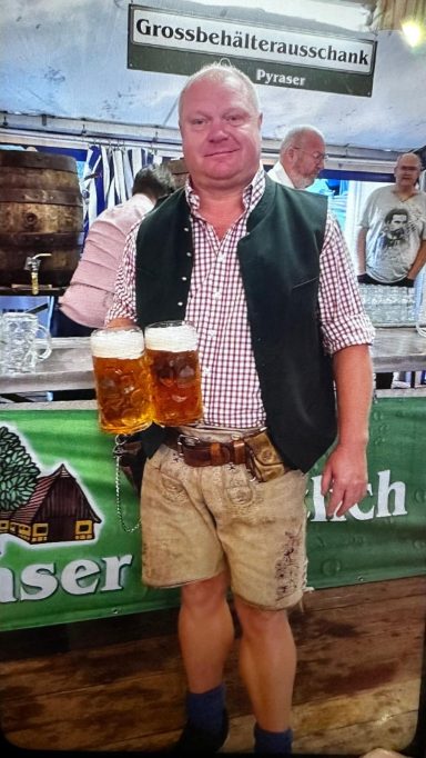 Mann in Tracht hält zwei Bierkrüge vor einer Feststand.